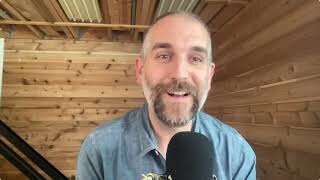 A man with a beard and mustache, wearing a blue shirt, speaks into a microphone in a wood-paneled room.