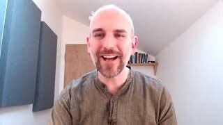 A man with a beard and short hair, wearing a grey shirt, smiles at the camera in a bright room with a wooden door, shelves, and wall panels.