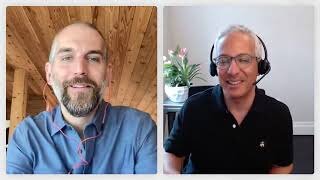 Two men are having a video call; one is in a wooden room wearing a blue shirt, the other wears glasses and a headset, sitting by a plant in a black shirt.