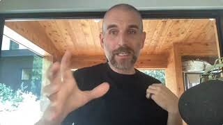 A man with a shaved head and beard, wearing a black shirt, gestures with his hand while speaking indoors near a window and wooden ceiling.