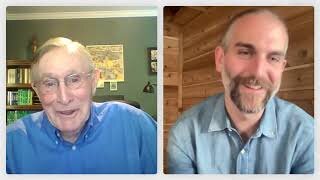Two men are having a video call; one is in a room with green walls and bookshelves, the other is in a room with wooden walls. Both are smiling and wearing blue shirts.