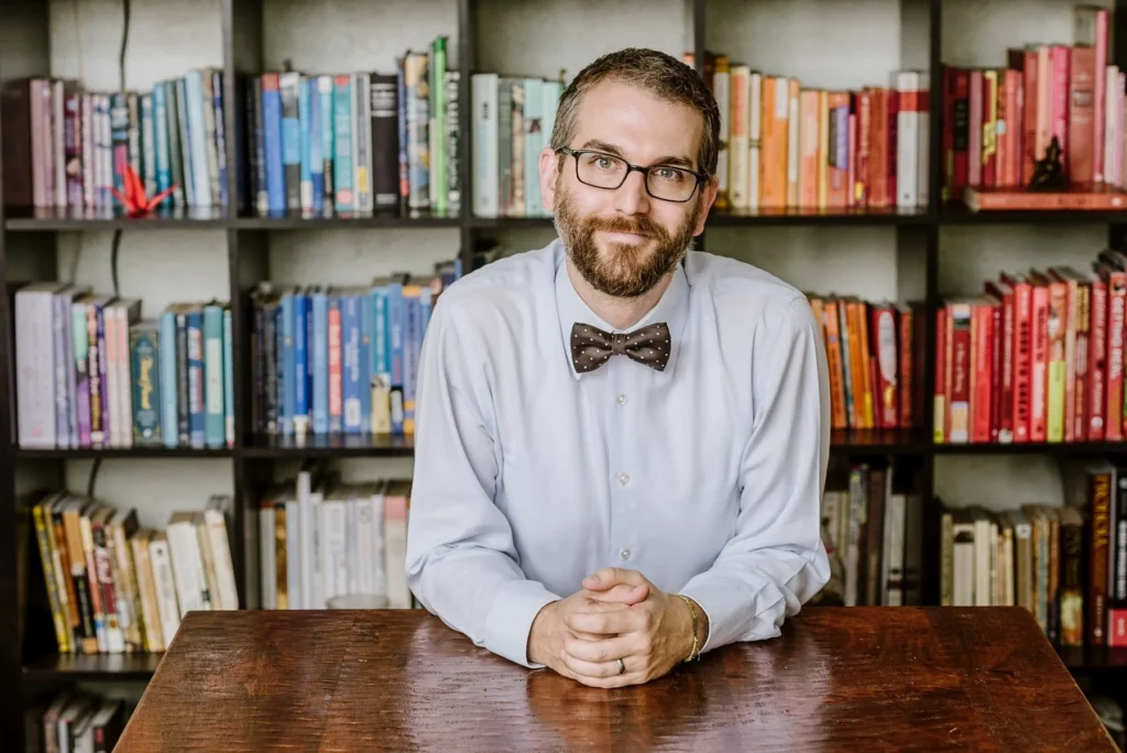 A person with glasses and a bow tie sits at a wooden table in front of bookshelves organized by color.