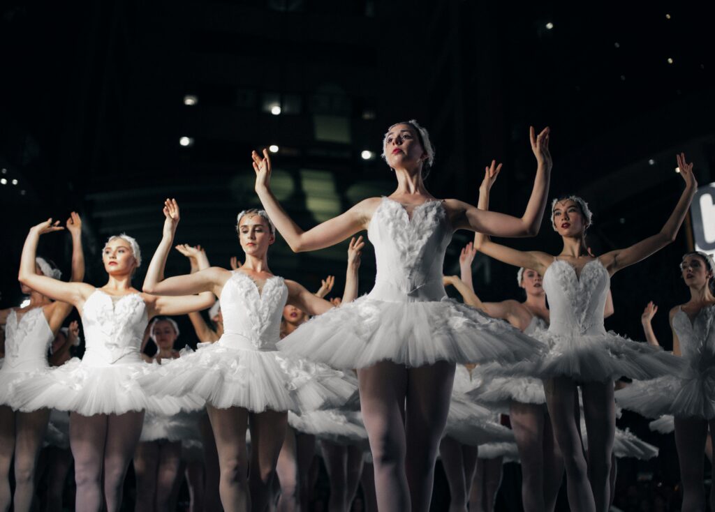 A group of ballerinas in white tutus perform on stage, holding their arms gracefully above their heads under stage lighting.