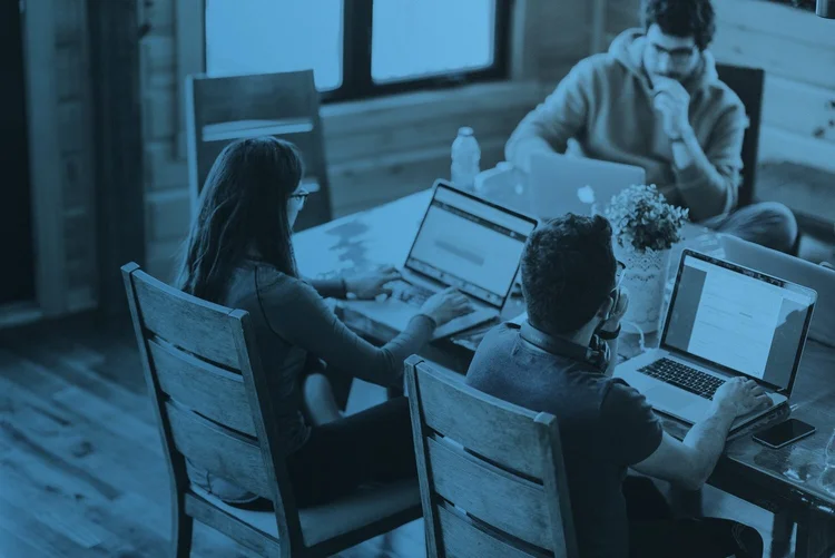 Three people sit at a wooden table working on laptops in a room with wooden floors and a window.