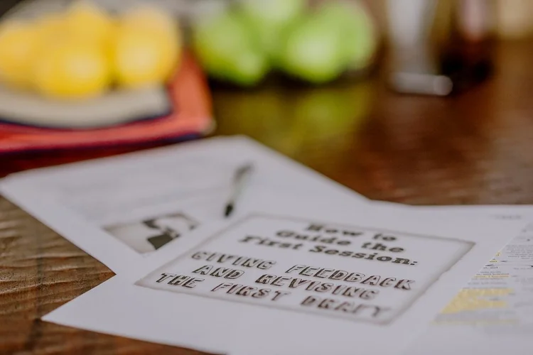 A close-up of printed papers on a table, one titled “Giving Feedback and Revising the First Draft,” with blurred lemons and apples in the background.