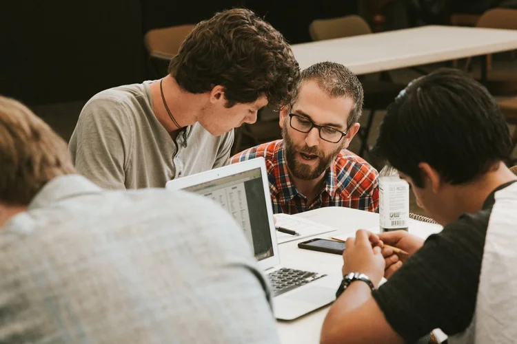 Four people sit around a table with laptops and papers, appearing engaged in a discussion or collaborative work in an indoor setting.