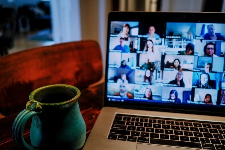A laptop displaying a video conference with many participants on screen sits on a wooden table next to a green coffee mug.