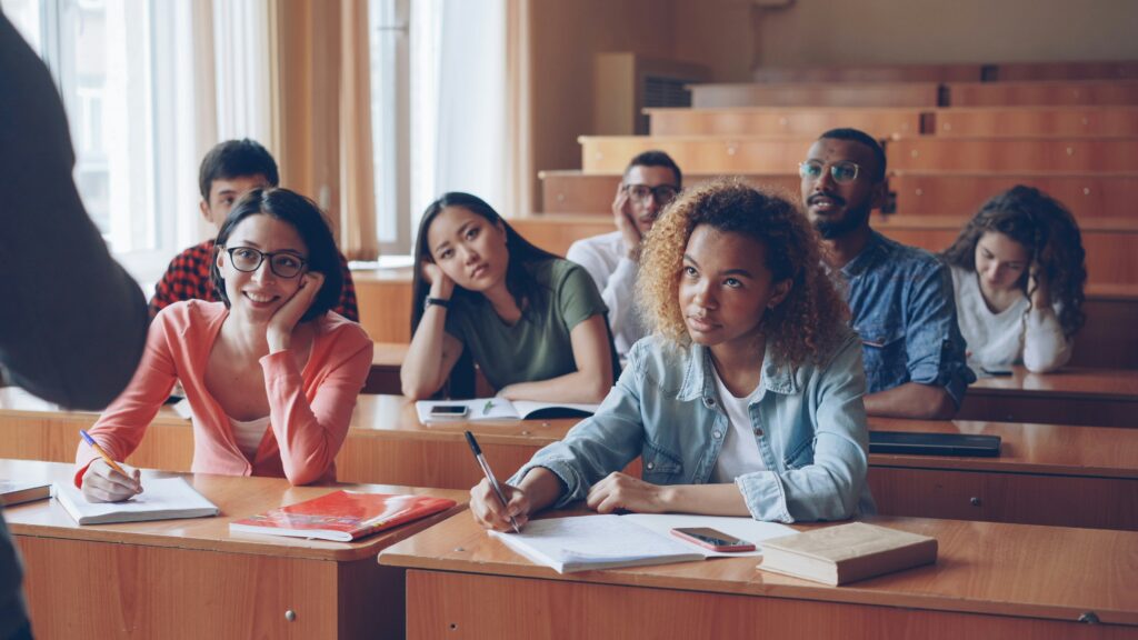 A group of students sit at desks in a classroom, listening to a person at the front while taking notes and looking attentive.
