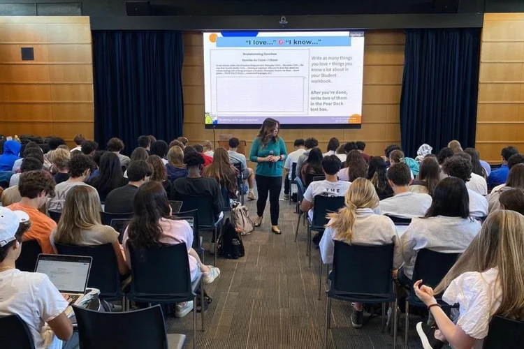 A teacher stands in front of a classroom filled with students, who are seated and facing a large screen displaying a presentation.