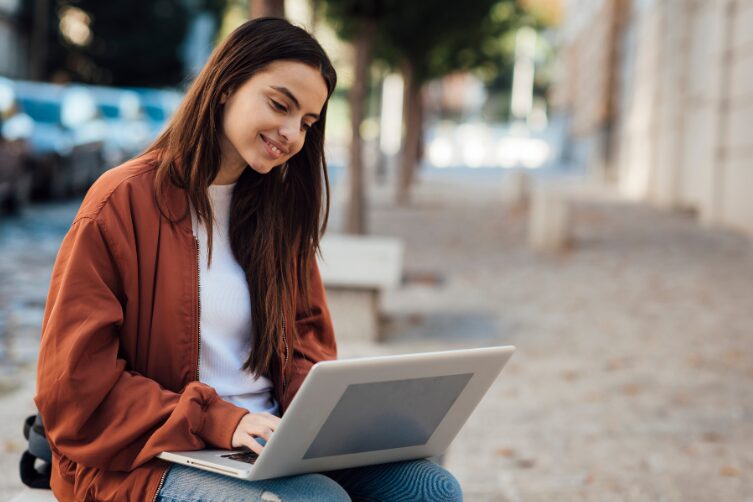 A young woman sits outdoors on a bench, smiling while using a laptop on her lap.