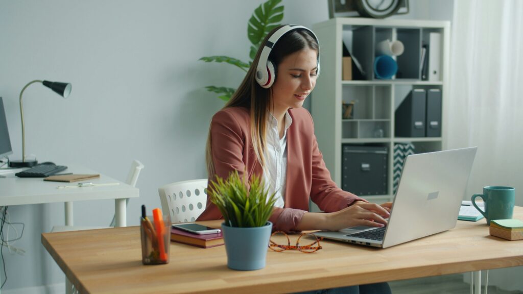 Woman wearing headphones works on a laptop at a desk with a plant, notebooks, and glasses in a modern home office setting.