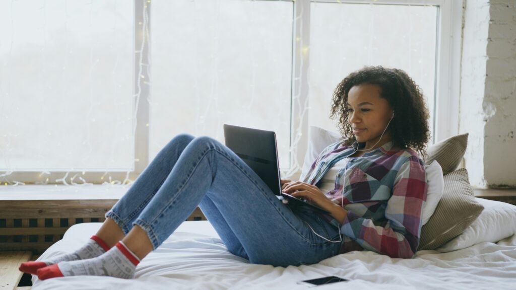 Woman wearing headphones sits on a bed and uses a laptop, with a smartphone lying beside her and large windows in the background.