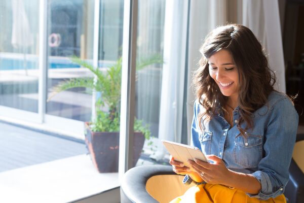Woman sitting indoors by a large window, smiling while using a tablet. A potted plant and bright sunlight are visible in the background.