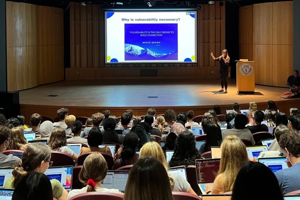 A speaker presents on vulnerability to an audience in an auditorium; the slide quotes Brené Brown and attendees use laptops.