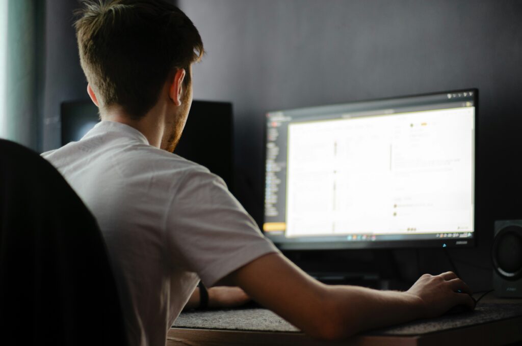 A person in a white shirt sits at a desk using a computer with a large monitor displaying a bright screen in a dimly lit room.