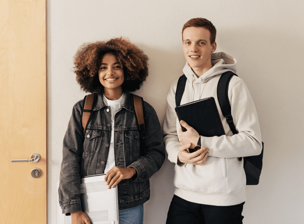 Two students with backpacks stand against a wall, smiling. One holds a laptop, the other holds a notebook.