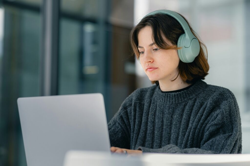 A person wearing headphones and a dark sweater sits at a desk, looking at a laptop screen in a bright indoor setting.