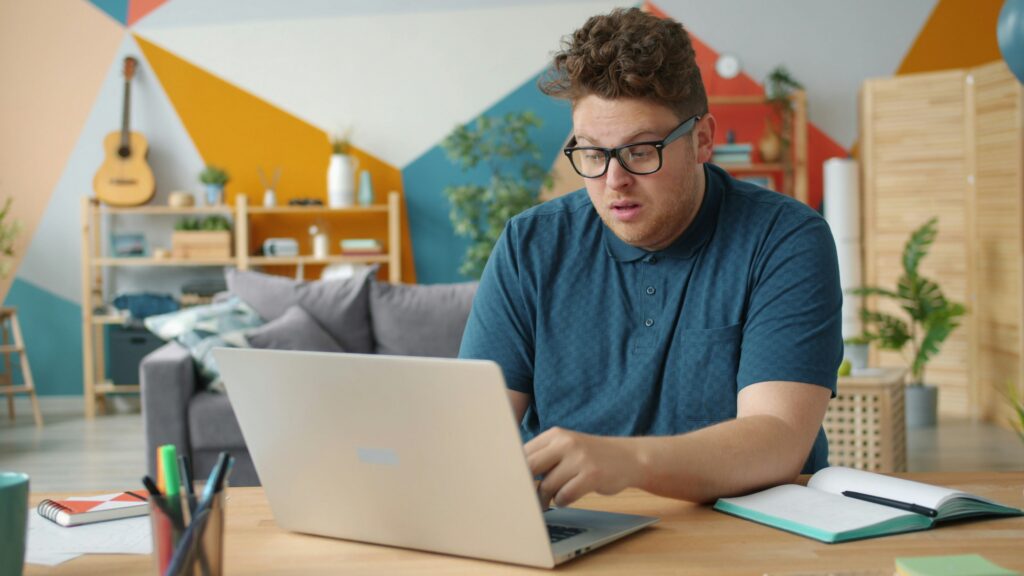 A person wearing glasses sits at a desk using a laptop, with an open notebook and a pen beside them in a colorful, modern living room.