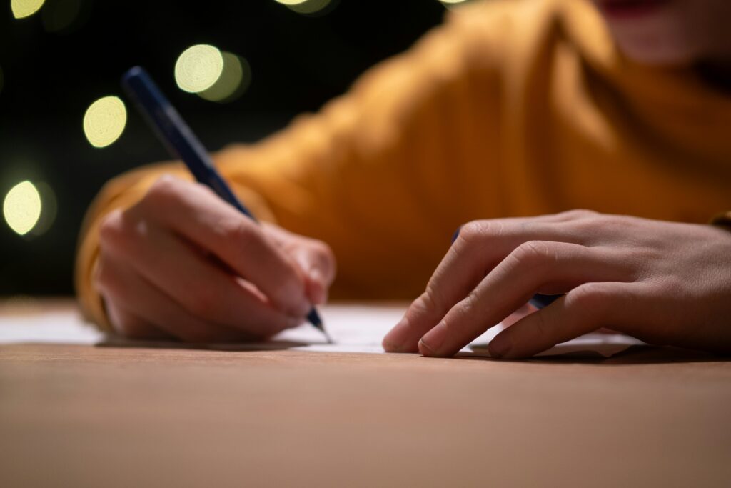 A person wearing a yellow sweater writes on paper with a pen at a table, with blurred lights in the background.
