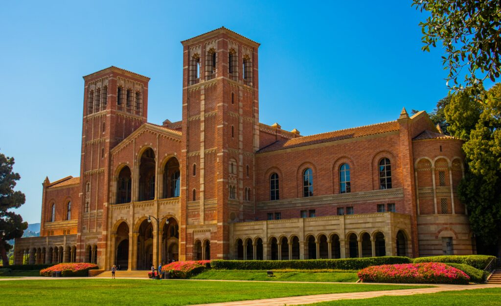 A large brick building with two towers, arched entrances, and landscaped grounds under a clear blue sky.