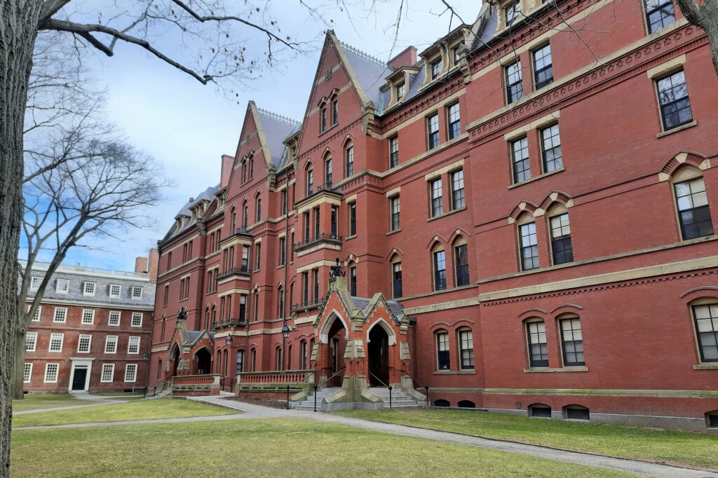 A large, historic red brick building with pointed roofs and arched doorways stands on a grassy courtyard with leafless trees.