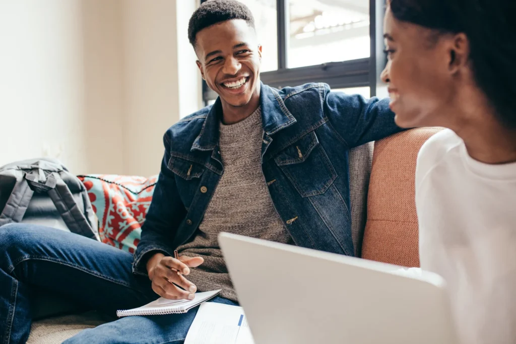 Two people sit on a couch, smiling and talking. One holds a notebook and pen, while the other uses a laptop.