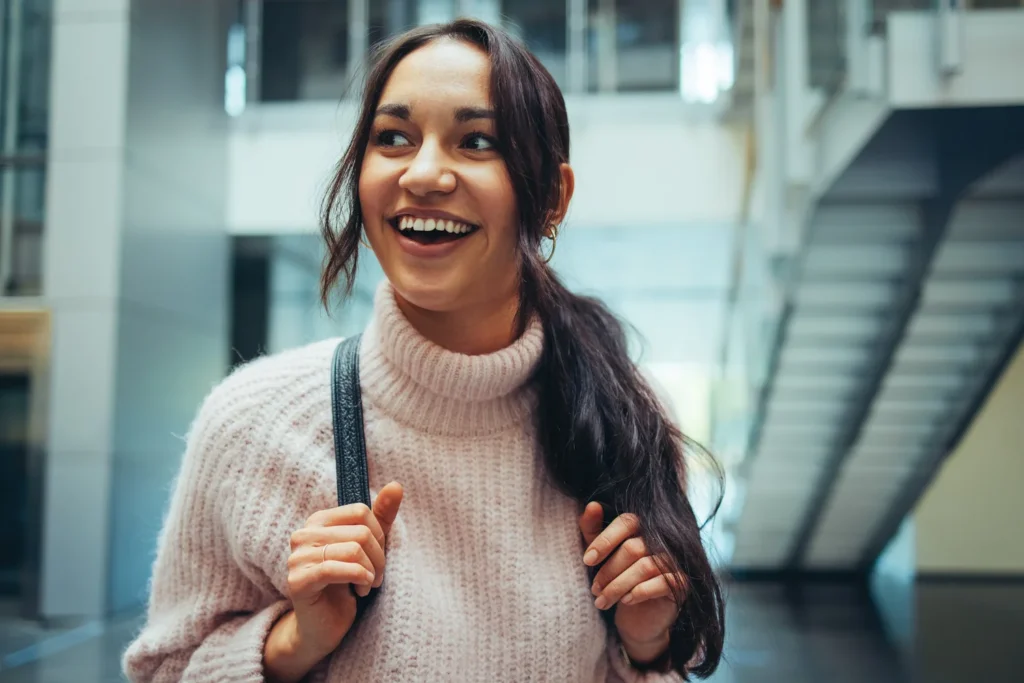A young woman wearing a light pink sweater smiles while holding the straps of her backpack in a modern indoor setting with stairs in the background.