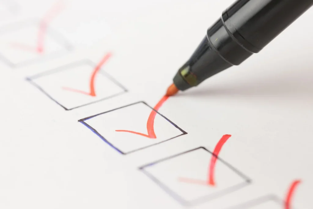 A black marker draws a red check mark in a square checkbox on a checklist, with other boxes already checked off in red.