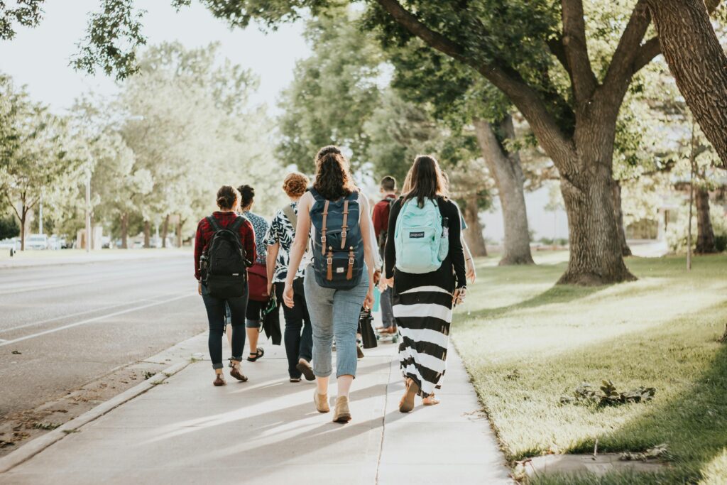 A group of people with backpacks walk along a sidewalk bordered by grass and trees on a sunny day.