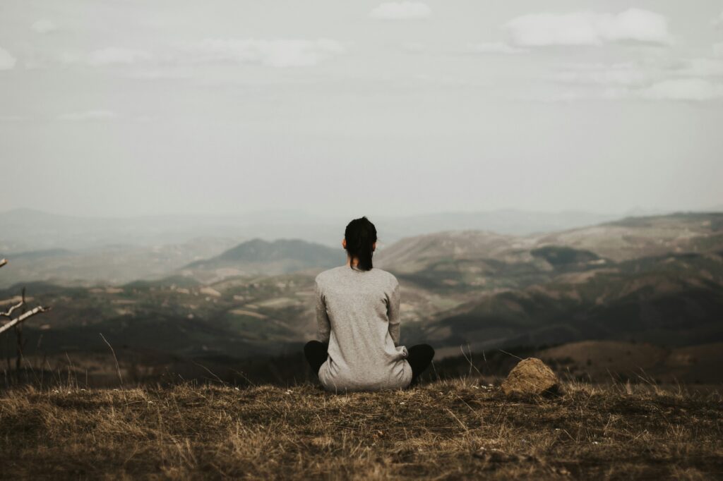 A person sits cross-legged on dry grass, facing distant mountains and a cloudy sky. The landscape appears vast and serene.