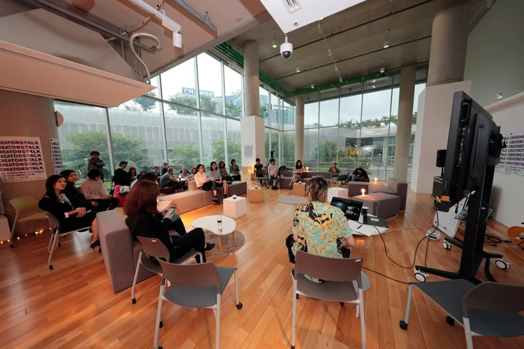 People seated in a modern, glass-walled room participate in a small group discussion or presentation, with a speaker facing an audience and a large monitor nearby.