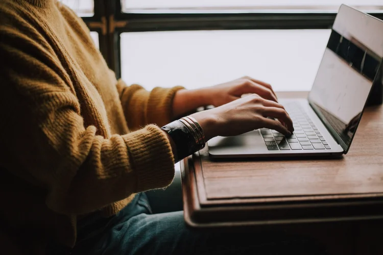 Person wearing a mustard sweater types on a laptop at a wooden table near a window.