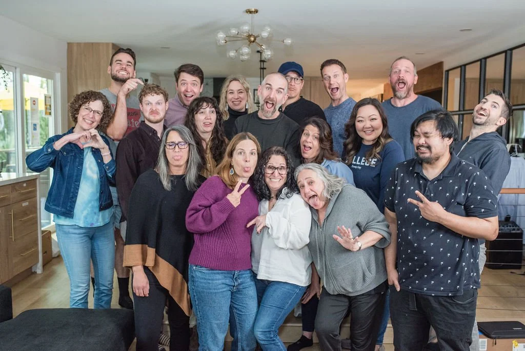 A group of sixteen adults poses indoors, standing close together and making playful faces and gestures for the camera.