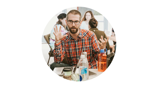 A man in a plaid shirt sits at a table with a laptop and water bottles, gesturing with both hands. Other people are visible in the background.